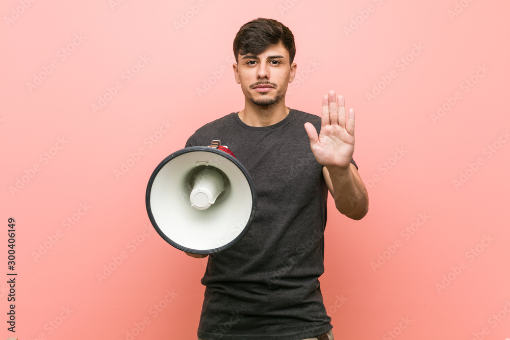 Wall mural young hispanic man holding a megaphone standing with outstretched hand showing stop sign, preventing