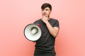 Young hispanic man holding a megaphone looking sideways with doubtful and skeptical expression.
