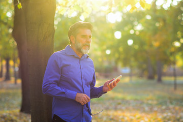 Happy smiling businessman with gray beard wearing blue shirt and using modern smartphone near tree in the park morning, successful employer to make a deal while standing near tree 