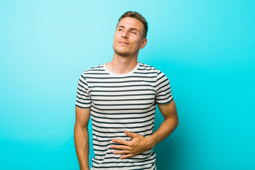 Young caucasian man against a blue wall touches tummy, smiles gently, eating and satisfaction...