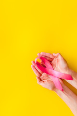Pink ribbon in hands as symbol of breast cancer awareness on yellow background top view copy space