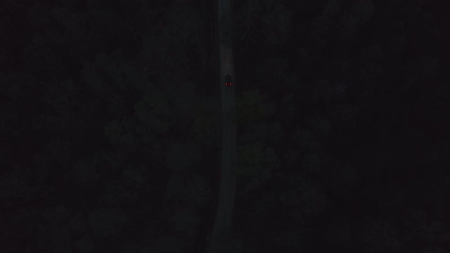 Aerial front view black car driving on dark countryside asphalt road among summer forest at night. View from drone car with headlights riding on night road through dark trees