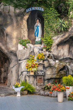 Da Nang, Vietnam - March 10, 2019: Closeup of Holy Virgin Mary in grotto outside Cathedral. Gray stones and green foliage with colored flowers in front under slogan: Ave Maria.