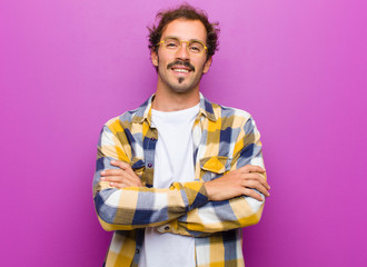 young handsome man looking like a happy, proud and satisfied achiever smiling with arms crossed against purple wall
