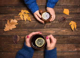 Mother and little daughter hands. Closeup. Hand holding a cup of tea on the wood table covered with autumn leaves. Mom and daughter drinking tea with lemon cold autumn day.