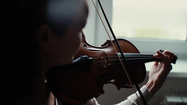 Close-up portrait of young woman musician playing the violin by the window in slow motion. Lovely girl is practicing playing musical instrument at home.