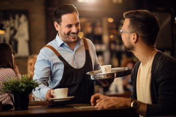 Happy waiter talking to a man while serving him coffee in a cafe.