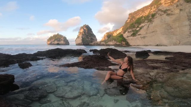A young, brunette woman with long hair in a black bikini taking a selfie photo with her smartphone, sitting and posing in the crystal clear water by herself alone.