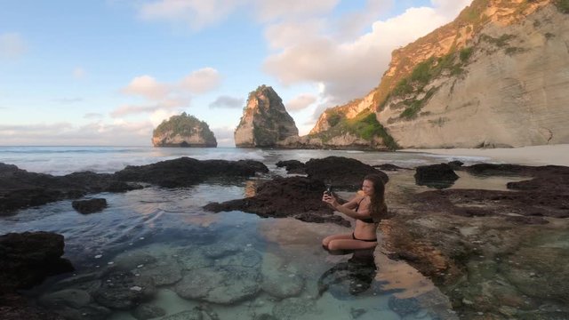 A young, brunette woman with long hair in a black bikini taking a photo of the beautiful view with her smartphone, sitting in the crystal clear water by herself alone.