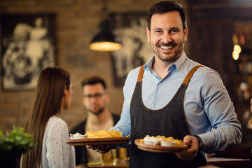 Happy waiter holding plates with food and looking at camera in a pub.