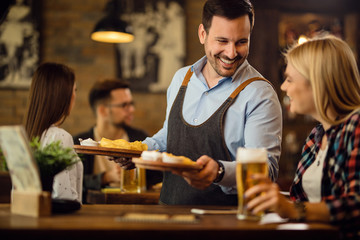 Happy waiter giving nachos to a woman who is drinking beer in a pub.