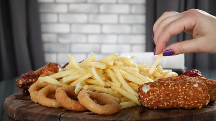 Wooden board with french fries and various beer snacks in the pub. Girl's hand takes food from a common large plate. A high-calorie snack for group friends. Tasty snacks. Beer snack.