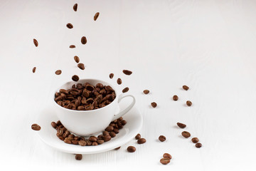 A cup with coffee beans in the air flying in flight like a spray. On background of a white wooden table, coffee shop advertising concept, copy space.