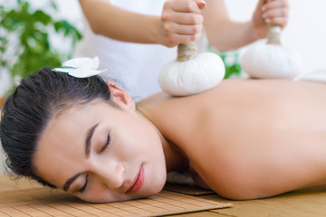 Obraz na płótnie Canvas Young pleased woman is getting thai massage, therapy. Female master is massaging client with herbal bags. Brunette girl is lying on couch in light spa ayurveda salon. Relax and health care concept.