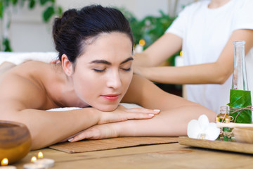 Fototapeta na wymiar Young pleased woman is getting thai massage, therapy. Female hands of master are kneading back of client. Brunette girl is lying on couch in light spa ayurveda salon. Relax and health care concept.