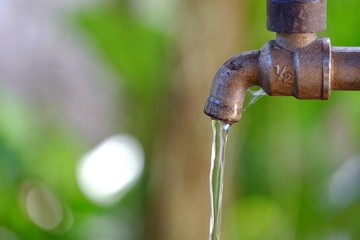 Water flowing from old steel faucet in outdoor space with blur green leaves background for saving water concept