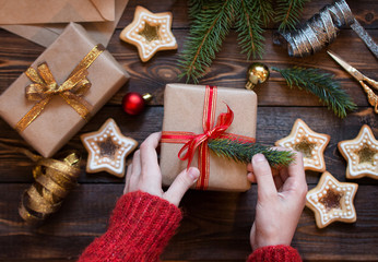 Womens hands pack Christmas gift on a wooden table with spruce branches, home-made cookies and Christmas tree toys. 