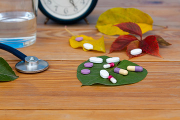 Autumn colds. Time to take pills. Alarm clock, stethoscope, colorful pills, autumn leaves of different colors on a wooden table.