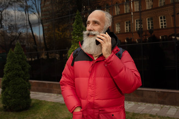 Handsome  senior bearded man is talking by a smartphone while having coffee break on a street beside office building. 