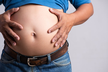 Naklejka premium Overweight or fat adult man hand holding belly stomach in very tight jeans on a gray background. Healthcare, medicine concept.