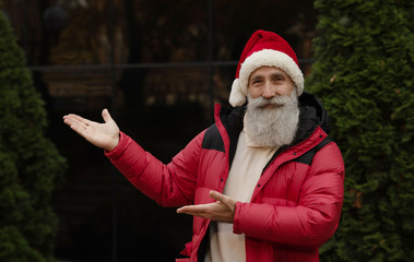 Obraz premium Senior man with beard in red santa hat presenting something outdoor. Xmas. New Year.