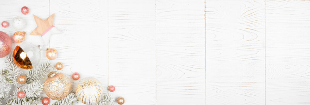 Christmas corner border banner of snowy branches and dusty rose, gold, and white ornaments. Overhead view on a white wood background.