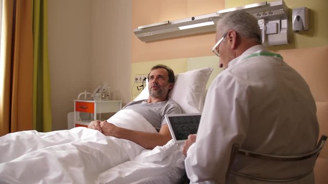 Adult ill man lying in hospital ward communicating with aging physician in white robe. Grey-haired practitioner watching brain scans on tablet and telling happy patient good news about MRI results