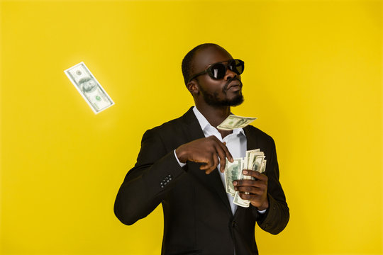 bearded solid young afroamerican guy is throwing out dollars from one hand, wearing sunglasses and black suit on the yellow background