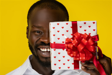 frontview of laughing bearded young afroamerican guy with one present in hand that closed half of face in white shirt  on the yellow background