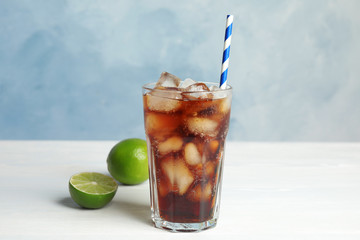 Glass of refreshing soda drink with ice cubes, limes and straw on white wooden table against blue background