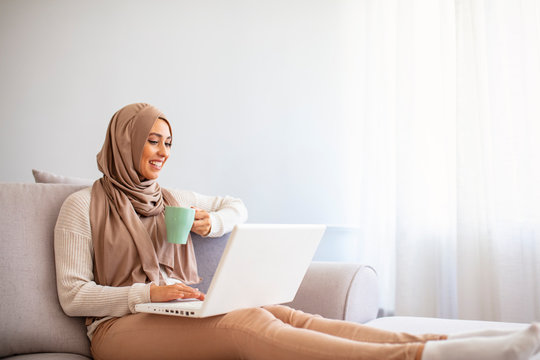 Happy muslim woman wearing hijab at home. Girl drinking coffee and shopping online on laptop, copy space. Shot of a young businesswoman using a laptop and having coffee at home.