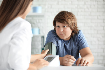 Female doctor checking overweight boy's blood pressure in clinic - obrazy, fototapety, plakaty
