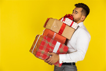 handsome young european guy is holding heavy packed gifts and presents on the yellow background