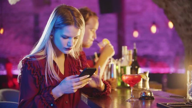 Young blonde woman sitting by the bartender stand and using her phone