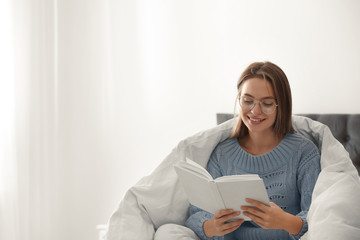 Young woman in warm sweater reading book on bed at home. Space for text