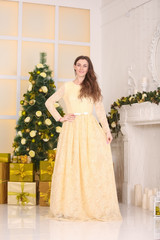 beautiful slim caucasian adult woman in long evening yellow dress celebrates christmas indoors in her white room with decorated xmas tree