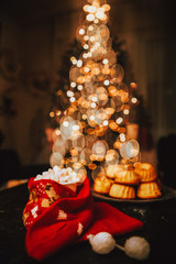 Obraz na płótnie Canvas Cup full of chocolate and small marshmellows bits wrapped up with a cristmas sock and a plate of hot mini bundt cake muffin, with a beautiful bokeh of a christmas tree light,