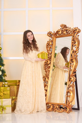 beautiful slim caucasian adult woman in long evening yellow dress holding gift box with bow and looking at the mirror while celebrating christmas in her white room