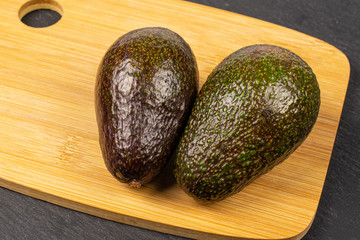 Group of two whole fresh green avocado on bamboo cutting board on grey stone