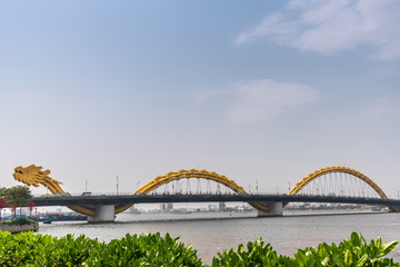 Da Nang, Vietnam - March 10, 2019: Shot from east bank over wide Han River on Cau Rong or Dragon bridge on horizon under morning sky. Green foliage in front.