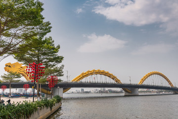Da Nang, Vietnam - March 10, 2019: Shot over wide Han River on head of Cau Rong or Dragon bridge between green foliage with red heart decoration under blue cloudscape.