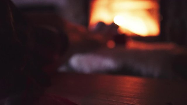 Feet in woollen socks by Burning Christmas Fireplace. 4K STABILIZED shot. Woman relaxes by warm fire with a cup of hot drink and warming up her feet. Close up. Winter and Christmas holidays concept.