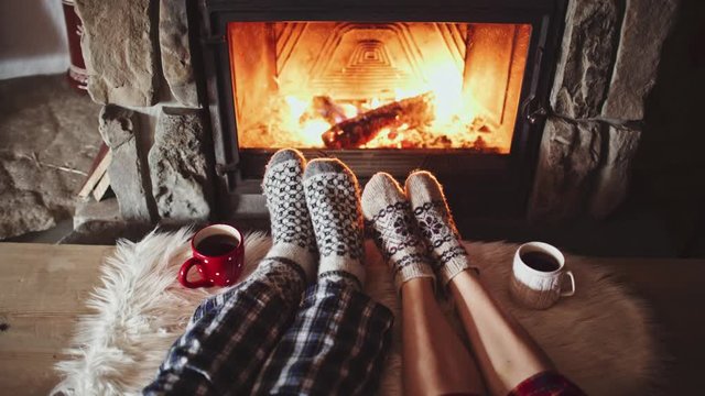 Couple Feet in Woollen Socks by the Cozy Fireplace, 4K. Man and Woman relax by warm fire and warming up their feet. Close up. Winter and Christmas holidays concept. 