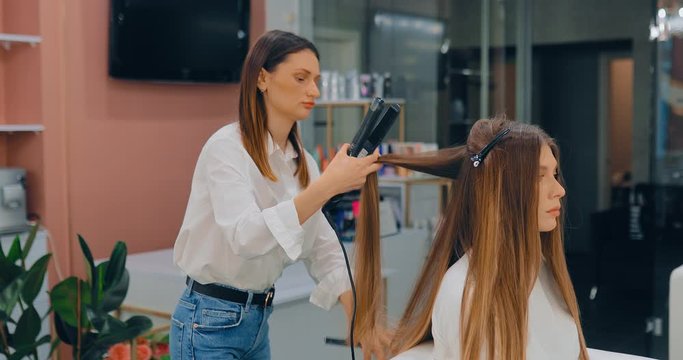 Professional hair straightening in a beauty salon. Beautiful young woman at a reception in a beauty salon, hairdresser straightens a lock of hair.