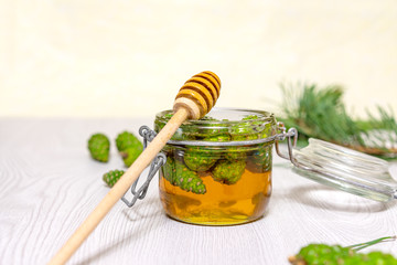Honey and young green pine cones in an open glass jar, and a spoon for honey on a light wooden table. Gourmet dessert. Natural medicine