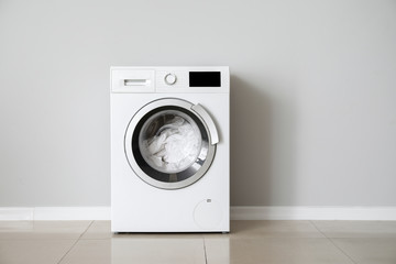 Modern washing machine with laundry near white wall