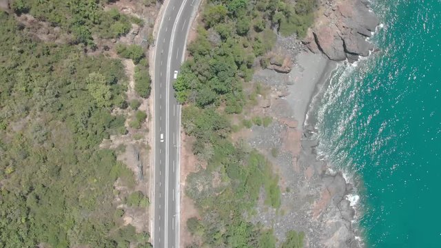 A flying camera takes a motorcyclist passing on a road near the sea with a view of the rocky shore.