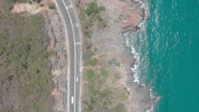 A flying camera takes a motorcyclist passing on a road near the sea with a view of the rocky shore.