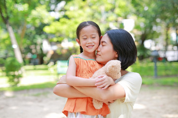 Portrait of happy Asian mother cuddle daughter and hugging teddy bear doll in the garden. Mom and child girl with love and relationship concept.