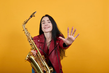 Portrait of crazy young woman in red casual clothes looking camera, keeping saxophone and smiling, standing with outstretched hands isolated on orange background. People lifestyle concept